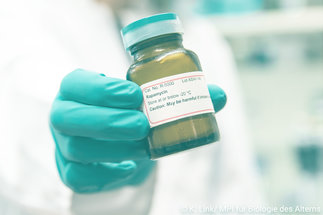 Eine Flasche mit einer Medizin wird von einem grünen Handschuh ins Bild gehalten. 