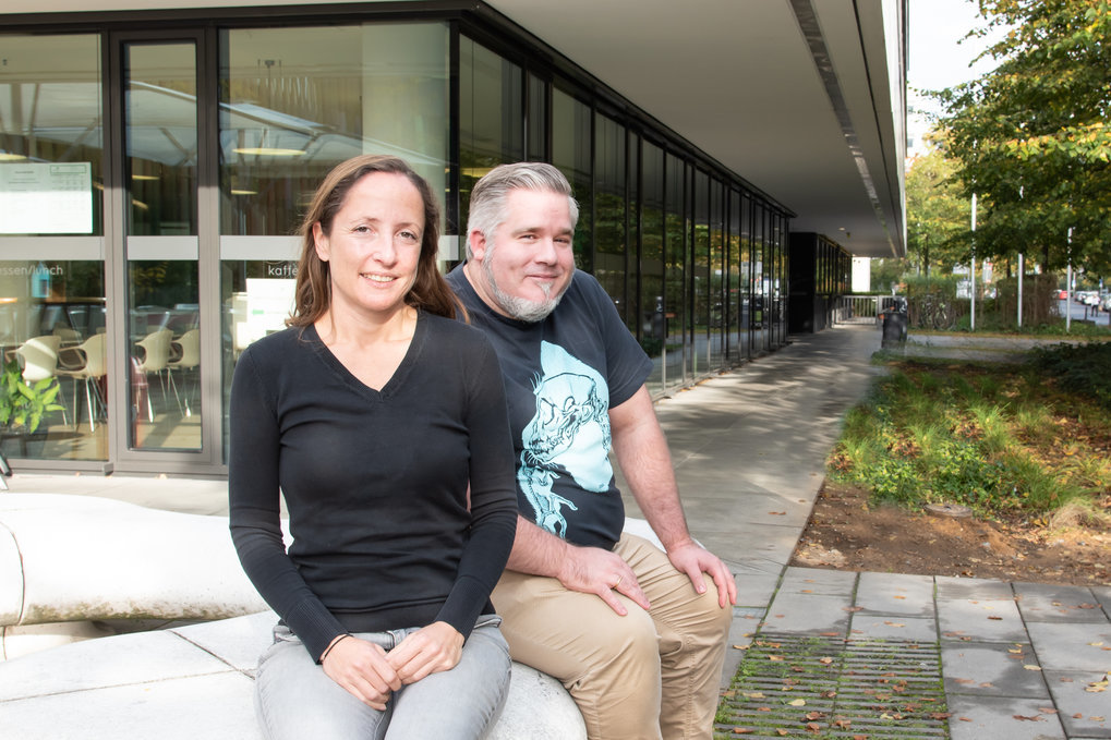 Two people sitting on a bench in front of a modern building with large windows and a landscaped area.