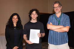 Three individuals, one holding a certificate, in a formal event setting with a dark backdrop.